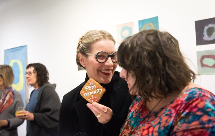 Curator Catherine Bell stands in the foreground alongside artist Eden Menta, who looks towards the cookie Catherine is holding in her hand which has the word 'FEM-AFFINITY' on it. Both are smiling at the opening of the 'FEM-AFFINITY' exhibition at Arts Project Australia in 2019, with artworks and people in the background.