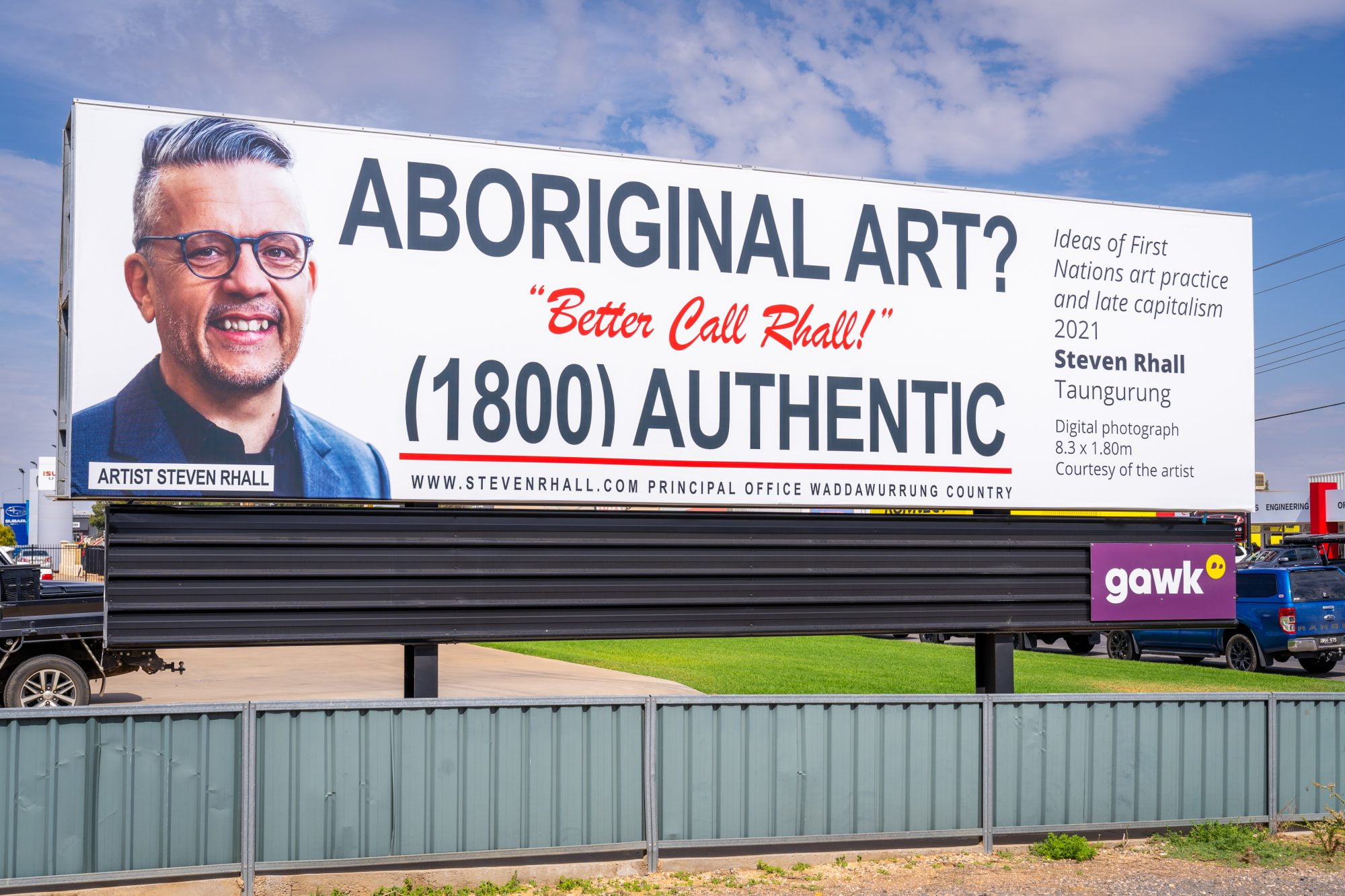 Image Description: ⁠
A billboard with black and red text, on the left is the head and shoulders of a smiling Taunarung man in a grey suit with overlaid text that reads “Artist Steven Rhall. The billboard's text reads: “Aboriginal Art? Better Call Rhall! (1800) Authentic.” The billboard is in an industrial area, the photograph  is taken from an angle high up (from a drone).