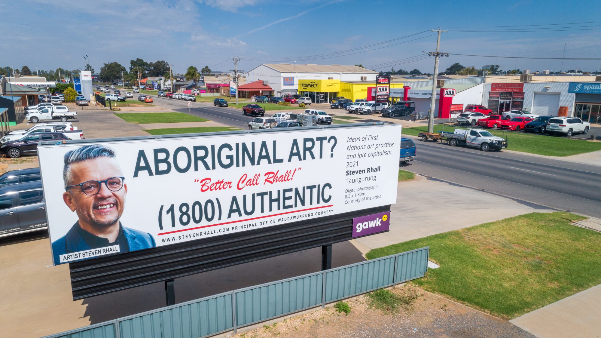 Image Description: ⁠
A billboard with black and red text, on the left is the head and shoulders of a smiling Taunarung man in a grey suit with overlaid text that reads “Artist Steven Rhall. The billboard's text reads: “Aboriginal Art? Better Call Rhall! (1800) Authentic.” The billboard is in an industrial area, the photograph  is taken from an angle high up (from a drone).