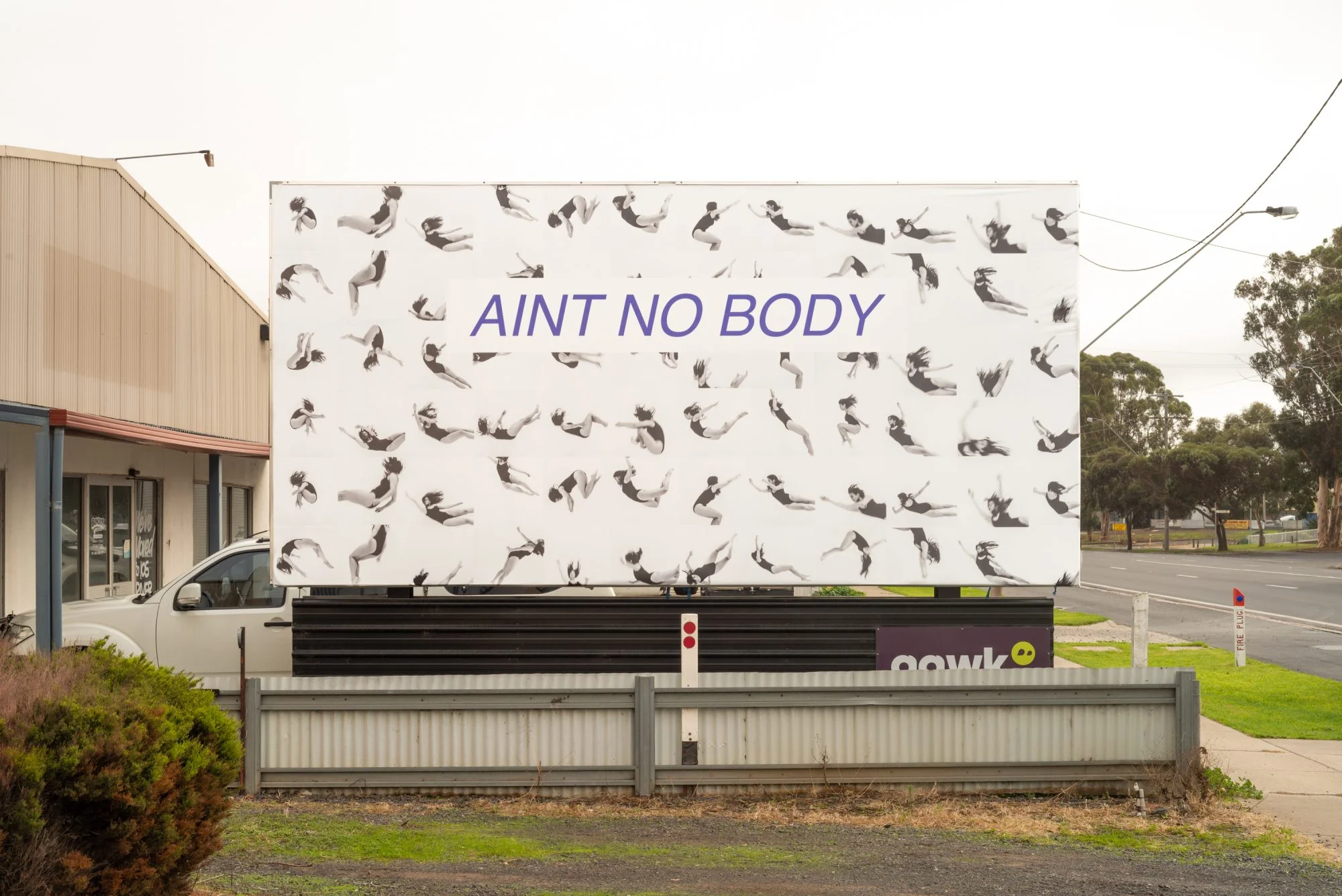 Image Description: A billboard with a black and white tiled images of the artist in a black leotard with long dark hair falling or jumping through the air, against a white background. Overlaid text reads ‘AINT NO BODY.’ The billboard is in an industrial area.