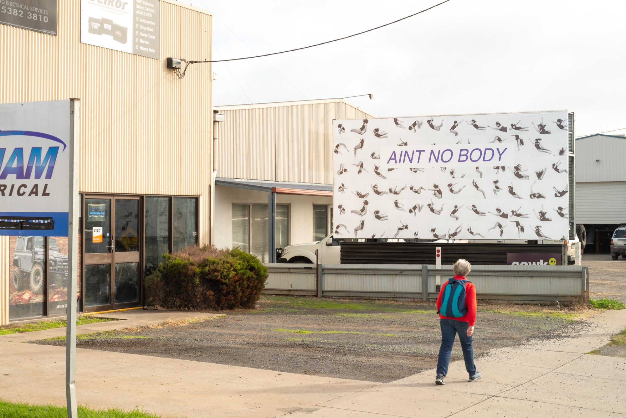 Image Description: A billboard with a black and white tiled images of the artist in a black leotard with long dark hair falling or jumping through the air, against a white background. Overlaid text reads ‘AINT NO BODY.’ The billboard is in an industrial area.