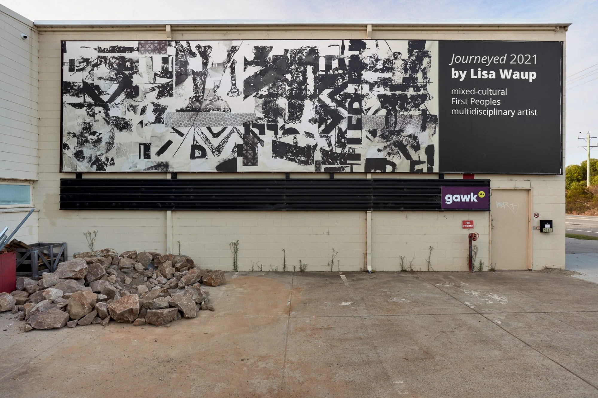 Image Description: A billboard filled collaged unintelligible black text and shapes on an off-white background. The billboard is installed the side of a building.